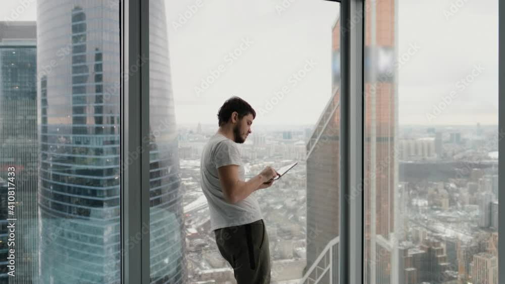 Young man with a tablet in his hands in a modern apartment near a window with a city view from the upper floor, writes text messages, looks through technologies Young man with a tablet in his hands in a modern apartment near a window with a city view from the upper floor, writes text messages, looks through technologies
