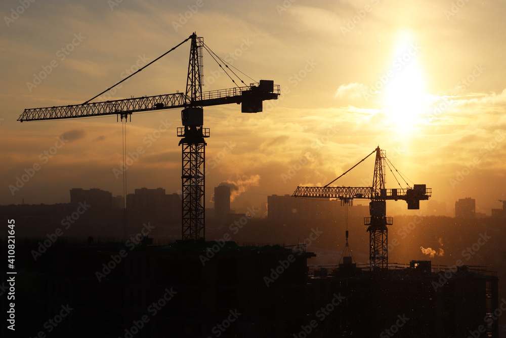 Silhouettes of construction cranes and unfinished residential building during snow on sunrise background. Housing construction, apartment block  in winter city
