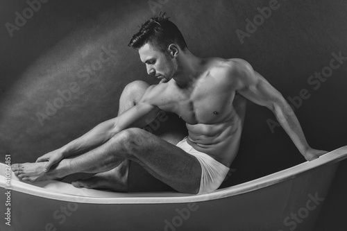 Young man in white panties with sexy bare muscular torso sitting on bathtub. Mans underpants and panties.