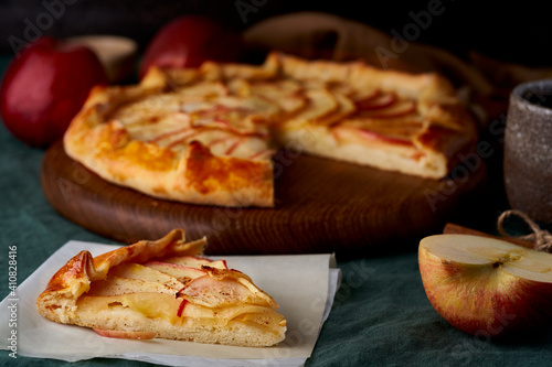 Apple pie with piece slice of galette with fruits, sliced sweet pastries on dark green tablecloth, sweet crostata on cutting wooden board, side view, autumn or winter food