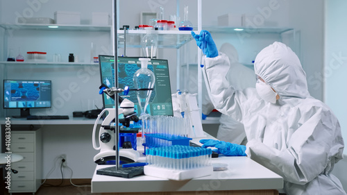 Biochemists in coverall typing at pc evolution of vaccine composition in equipped laboratory analysing test tube with sample. Doctors examining virus evolution using high tech researching diagnosis