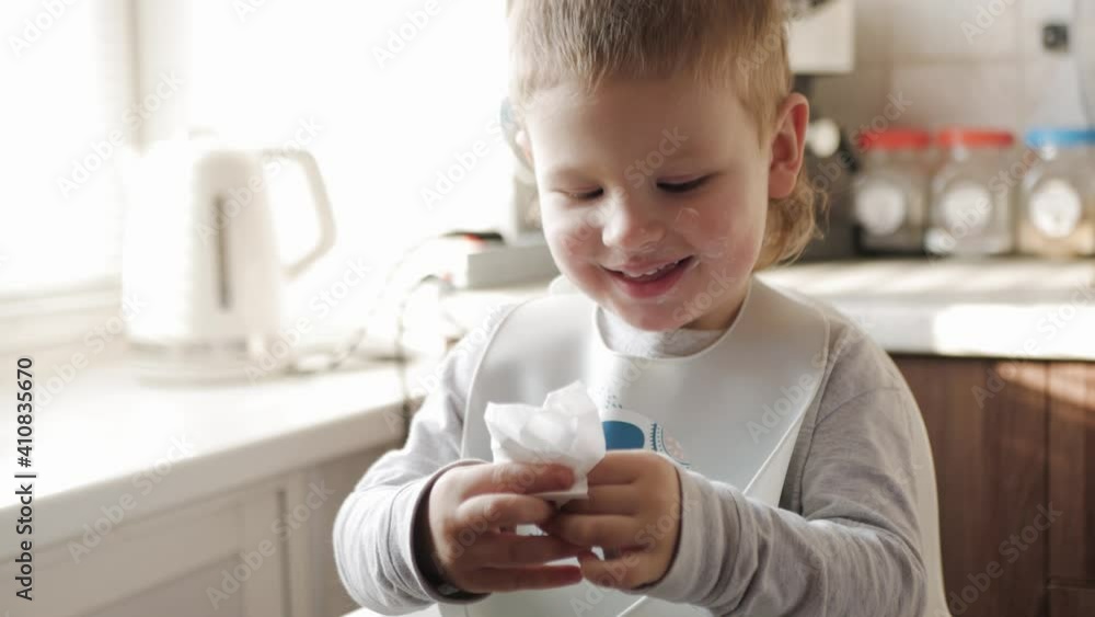 A small child sitting at the kitchen table wipes his grimy face after eating with a paper napkin. The boy smiles, tries to be clean, take care of himself. A small child sitting at the kitchen table wipes his grimy face after eating with a paper napkin. The boy smiles, tries to be clean, take care of himself.