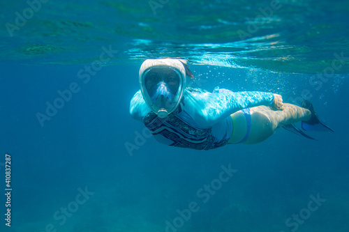 Woman swimming in blue water in full-face mask. Young woman swimming on sea surface. Snorkel in coral reef of tropical sea. Woman in snorkeling mask underwater photo. Blue water sport activity.