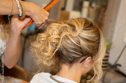 Woman young hairdresser making hairstyle to blonde bride girl in beauty salon