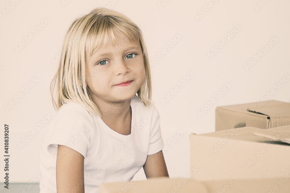 Positive cutest fair hair little girl sitting on floor near cartoon boxes in new apartment and looking inside. Family in new apartment concept Positive cutest fair hair little girl sitting on floor near cartoon boxes in new apartment and looking inside. Family in new apartment concept