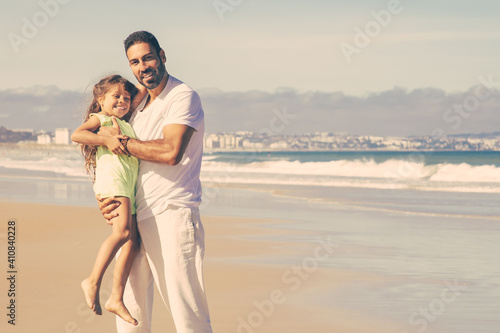 Joyful handsome dad holding happy little daughter in arms, standing wet sand, enjoying leisure time with girl on beach at sea. Family outdoors concept