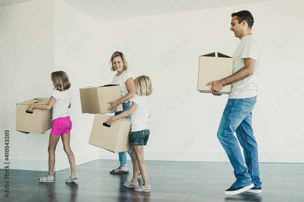 Happy family carrying cardboard boxes from empty room. Two little daughters helping parents with relocation in new apartment or house. Mortgage, relocation and moving day concept Happy family carrying cardboard boxes from empty room. Two little daughters helping parents with relocation in new apartment or house. Mortgage, relocation and moving day concept