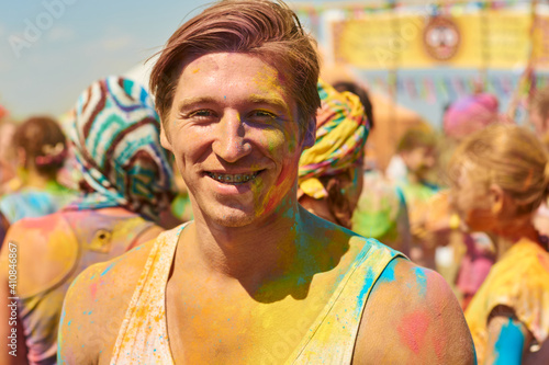 handsome young european athletic man with blond hair smeared in colorful paint on holi festival against the background of people in colored paint