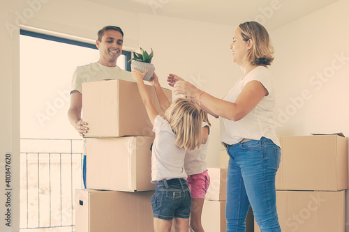 Happy young family with moving boxes in their new house. Mom, dad and children standing near cardboard boxes, smiling. Little girl holding plant in pot. Mortgage, relocation and moving day concept