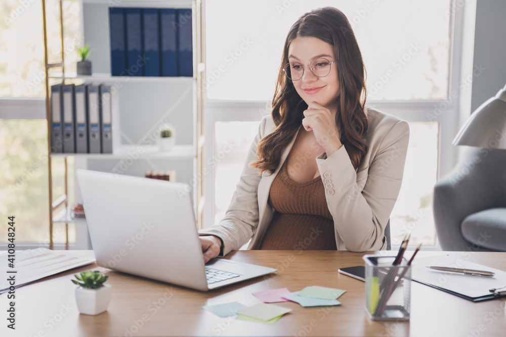 Photo of pregnant worker sit table laptop finger chin think wear glasses suit brown shirt workstation office indoors Photo of pregnant worker sit table laptop finger chin think wear glasses suit brown shirt workstation office indoors