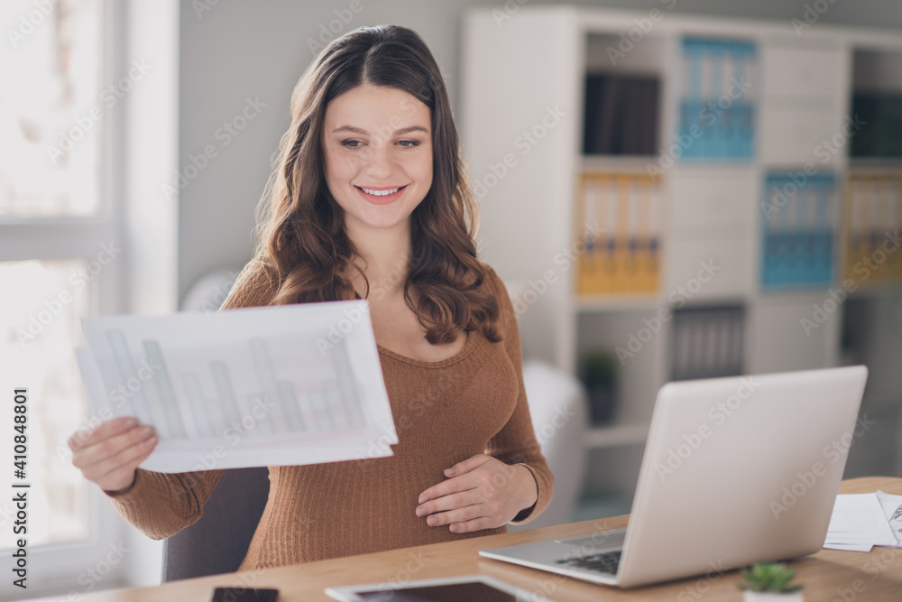 Photo of happy pregnant agent sit table hold paper sheet read wear brown shirt workstation office indoors Photo of happy pregnant agent sit table hold paper sheet read wear brown shirt workstation office indoors