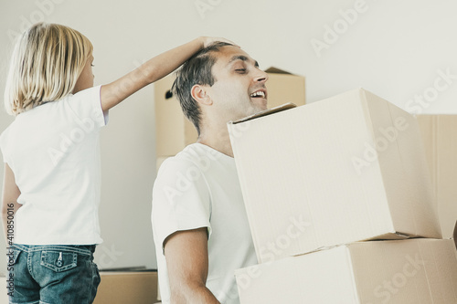 Middle-aged dad holding two carton boxes and sitting with closed eyes. Little girl touching father head. Mother talking with daughter. Family relocating. Mortgage, relocation and moving day concept