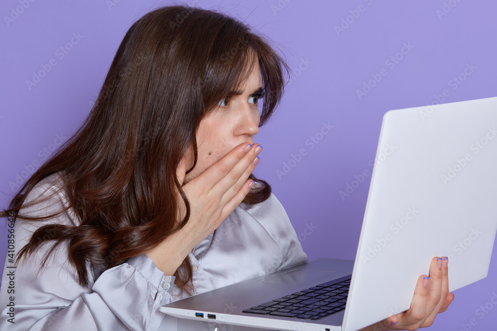 Amazed emotional woman with dark hair, looking at laptop screen with shocked expression, covering her opened mouth, isolated over lilac background, sees something bad, has trouble with work. Amazed emotional woman with dark hair, looking at laptop screen with shocked expression, covering her opened mouth, isolated over lilac background, sees something bad, has trouble with work.