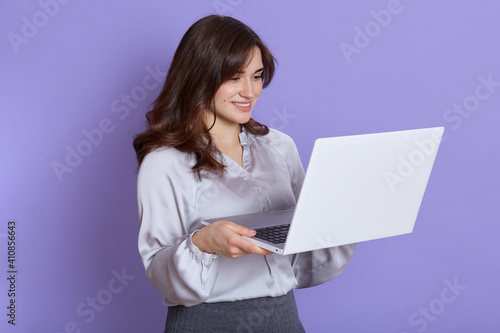 European young brunette woman girl in elegant white blouse and skirt posing isolated over lilac background, looking at screen with smile, winsome lady working online.