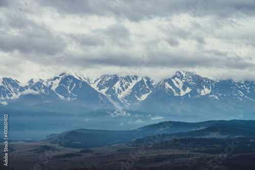 Scenic mountain landscape with great snowy mountain range among low clouds and green forest in valley at early morning. Atmospheric alpine scenery with blue white high mountain ridge under cloudy sky.