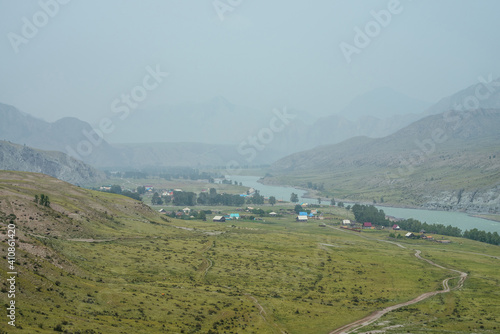 Scenic alpine landscape with mountain village by mountain river in mist. Atmospheric misty scenery with countryside on river bank. Beautiful view to mountain village on riverside in rainy weather.