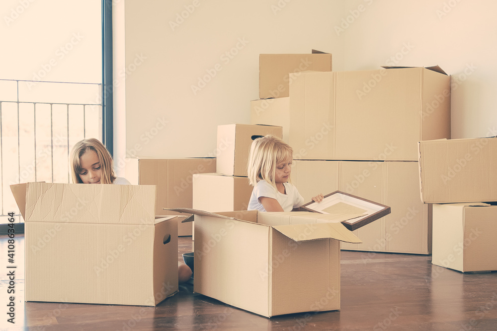 Focused little kids unpacking things in new apartment, sitting on floor and taking objects from open cartoon boxes. Relocation or moving concept Focused little kids unpacking things in new apartment, sitting on floor and taking objects from open cartoon boxes. Relocation or moving concept
