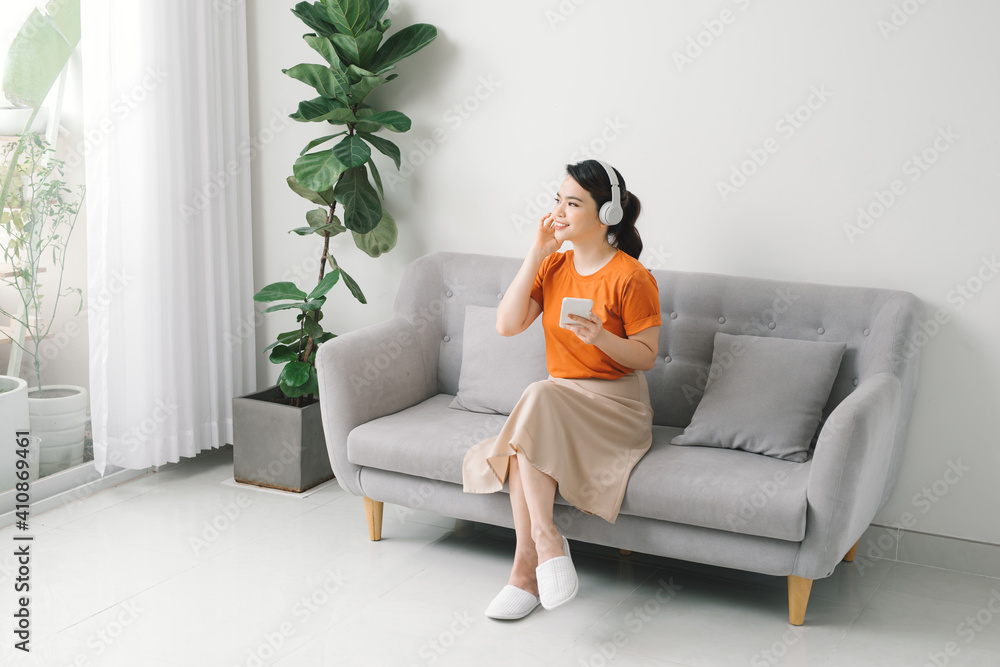Obraz premium Smiling girl relaxing at home, she is playing music using a smartphone and wearing white headphones