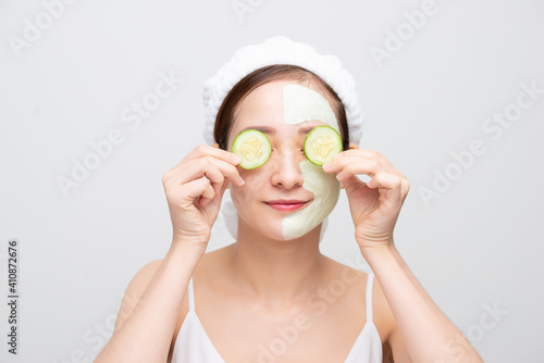 Beautiful young Asian woman with clay mask holding orange cucumber covering eye over white background.