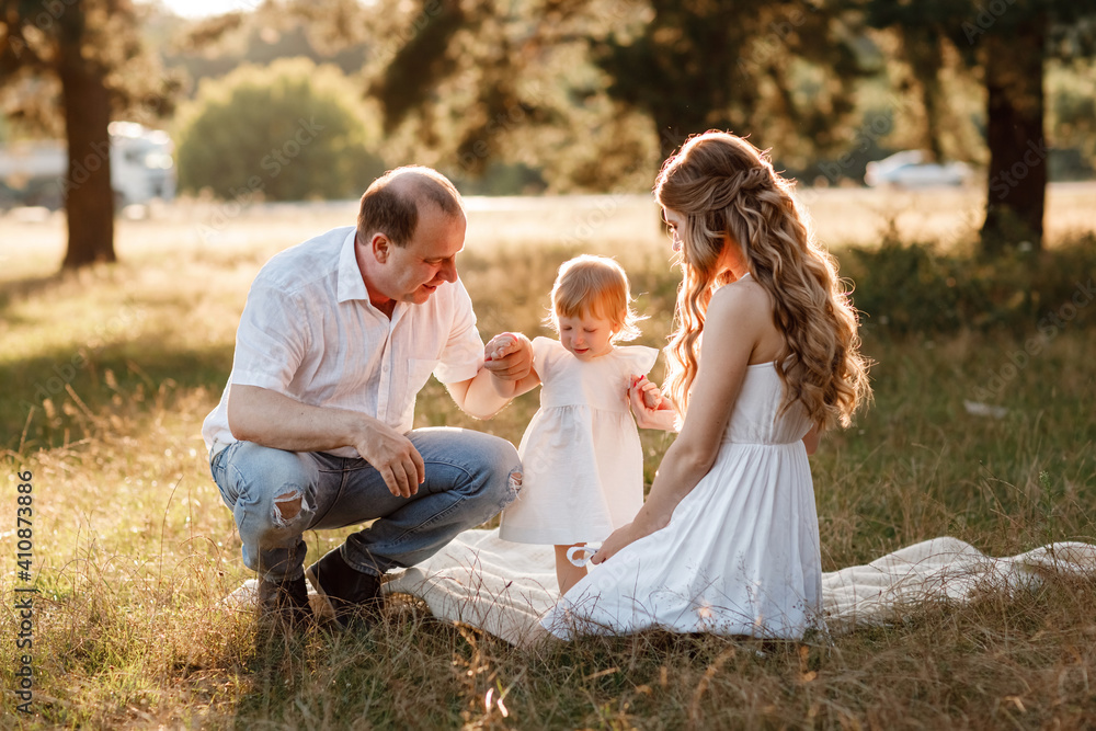 Fototapeta premium Smiling young family with little baby girl spending time together at the park, having a walk. Mom, dad and child playing outdoors at the sunset time. Concept of friendly family.