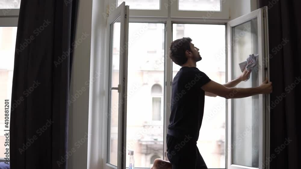 Houseman is cleaning window, cleans surface, uses a rag, standing in modern apartment Houseman is cleaning window, cleans surface, uses a rag, standing in modern apartment