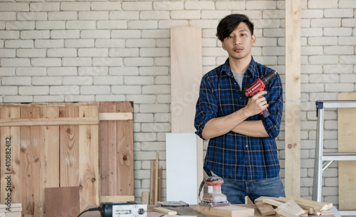 Good looking Asian carpenter standing while working for DIY jobs in carpenter room with several kinds of woods and types of equipment. He holding drill and cross his arms