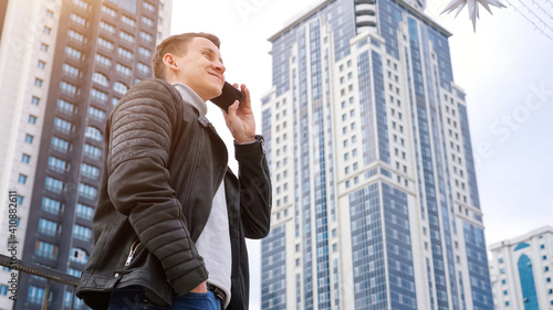 Confident businessman wearing autumn leather jacket speaks on modern smartphone standings against highrise buildings close low angle shot