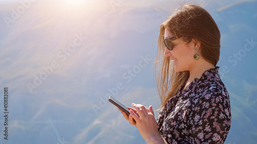 Young woman with phone against the background of mountains on a sunny day, hair fluttering in the wind.