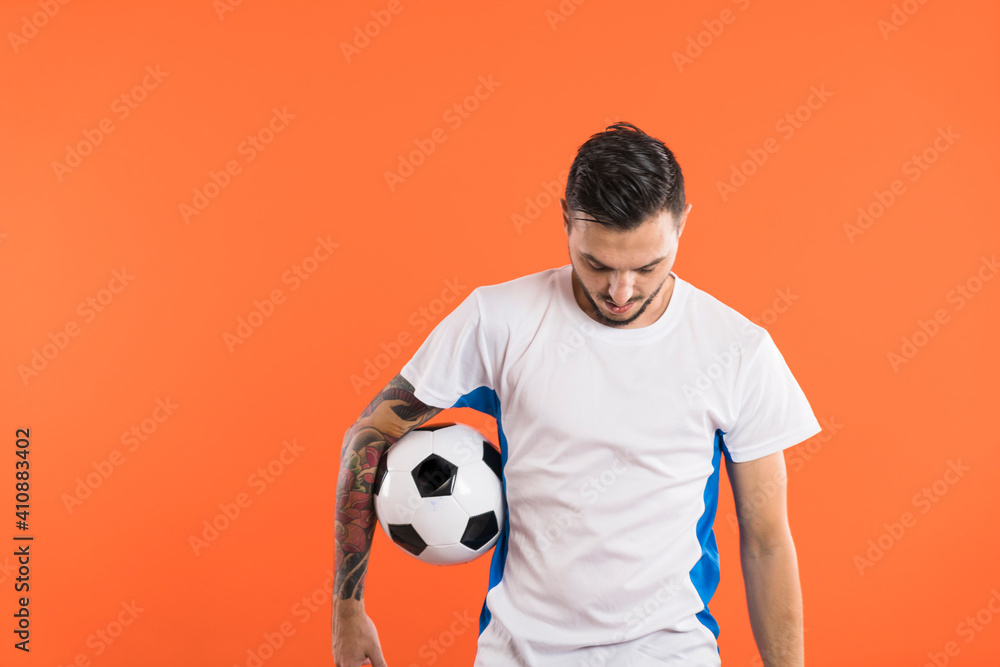 Soccer player looking down and holding a ball with one arm isolated on orange background Soccer player looking down and holding a ball with one arm isolated on orange background