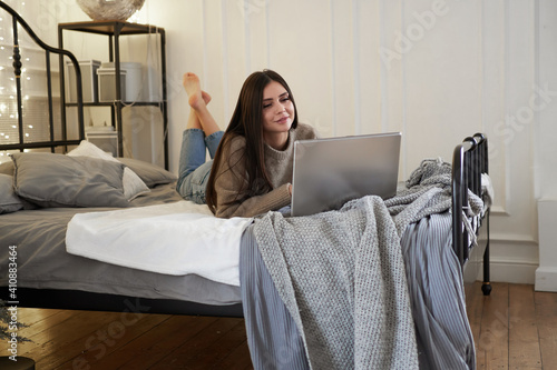 Young beautiful bruenette woman in cozy sweater in bed at home working on laptop photo