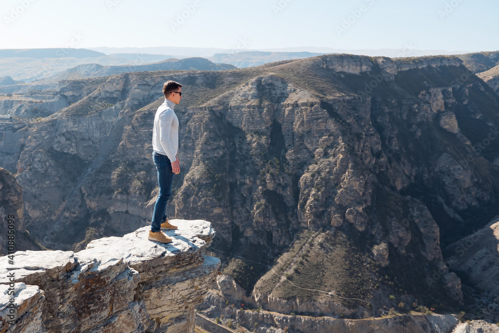 Courageous guy in sunglasses stands on dangerous steep cliff edge of grey colour against endless rocky hills under bright sunlight in autumn