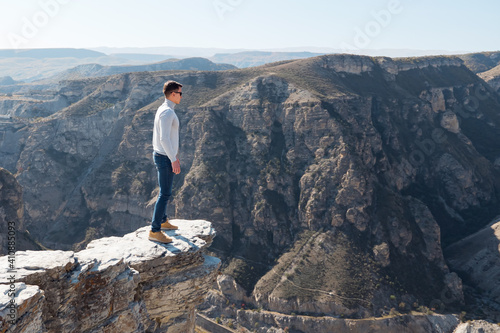 Courageous guy in sunglasses stands on dangerous steep cliff edge of grey colour against endless rocky hills under bright sunlight in autumn