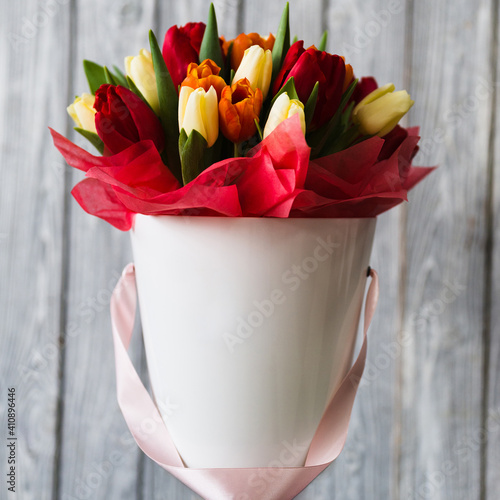 White flowerpot of red yellow orange tulips with red and pink ribbons on gray wooden background