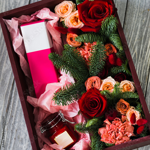 A burgundy wooden box with red roses, branches of a green Christmas tree, a pink perfume box in a glass jar lies on a wooden gray background