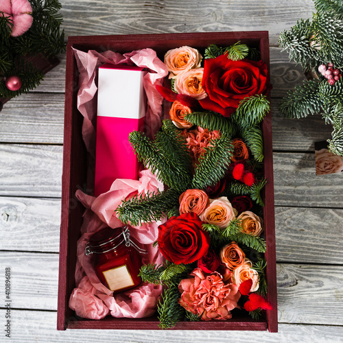 A burgundy wooden box lies on a gray wooden board inside which lies red roses with parts of the foliage of a Christmas tree with a pink perfume box and a glass jar