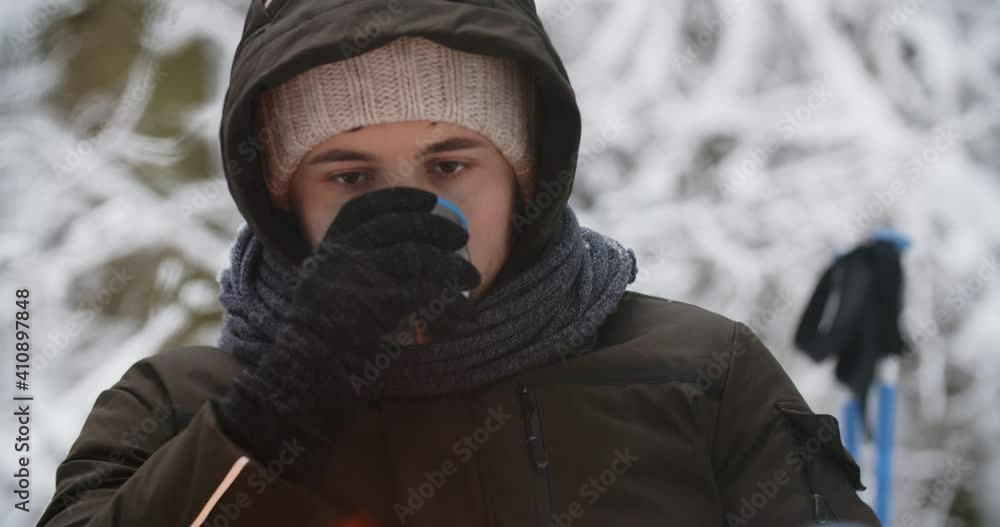 Portrait tired man sitting by campfire in snowy forest drinking hot tea. Young caucasian hiker warms up resting by burning fire after walk in winter woods enjoying nature. Human and nature concept Portrait tired man sitting by campfire in snowy forest drinking hot tea. Young caucasian hiker warms up resting by burning fire after walk in winter woods enjoying nature. Human and nature concept