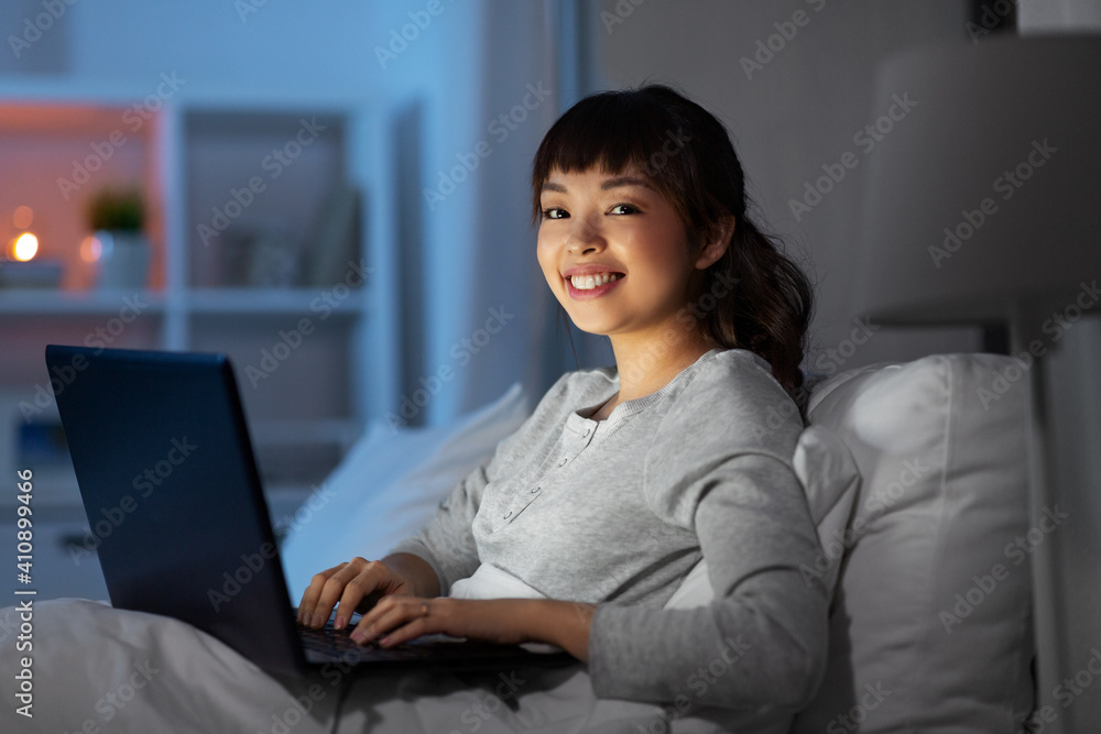 technology, internet, communication and people concept - happy smiling young asian woman with laptop computer lying in bed at home at night technology, internet, communication and people concept - happy smiling young asian woman with laptop computer lying in bed at home at night