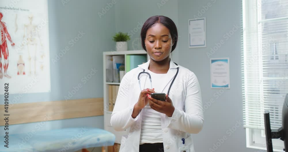 Close up of african american doctor therapist standing in office. Young woman professional looking at smartphone, phone, smiling. Portrait of physician, general practitioner in white coat. Close up of african american doctor therapist standing in office. Young woman professional looking at smartphone, phone, smiling. Portrait of physician, general practitioner in white coat.