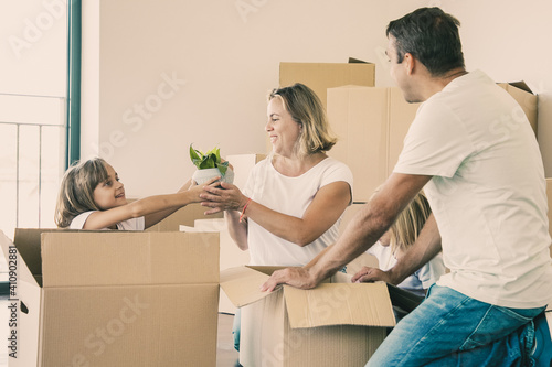 Cheerful parents and kids unpacking things in new apartment, sitting on floor and taking houseplant from open box. House buying or moving concept