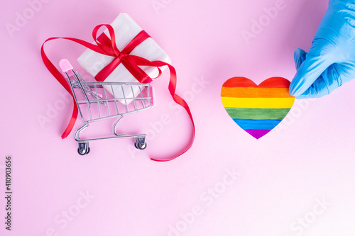 A hand in a protective blue glove holds a rainbow paper heart next to a gift in a shopping trolley on a pink background. Safe holiday greetings concept. Safe online shopping concept. LGBTQL concept photo