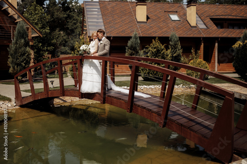 Beautiful wedding couple is standing on the wooden bridge. The bride in tulle veil and elegant hairdo is holding hands with her bearded groom in bow tie. Rustic outdoors stylish love story.