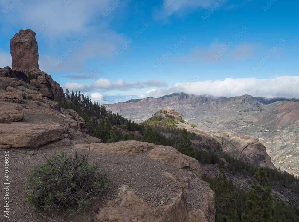 View of Roque Nublo rock formation in inland central mountains from famoust Gran Canaria hiking trail. Green pine trees and blue sky background. Canary Islands, Spain.