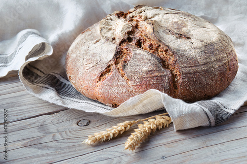 No knead handmade loaf on cutting board on wood with linen towel, spelt ears. German Bauernbrot means Rustic Farmers Bread in English. Wholemeal rye wheat bread baked in ceramic pan at home.