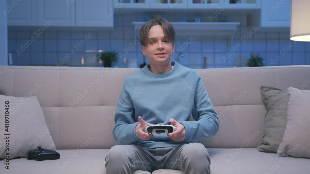 Portrait of Young Handsome Gamer Boy Sitting on Sofa and Win Playing in Video Games on Digital Console. Excited Player Guy Controlling Joystick for Videogame. One Person Focused on Gaming in Home Room Portrait of Young Handsome Gamer Boy Sitting on Sofa and Win Playing in Video Games on Digital Console. Excited Player Guy Controlling Joystick for Videogame. One Person Focused on Gaming in Home Room