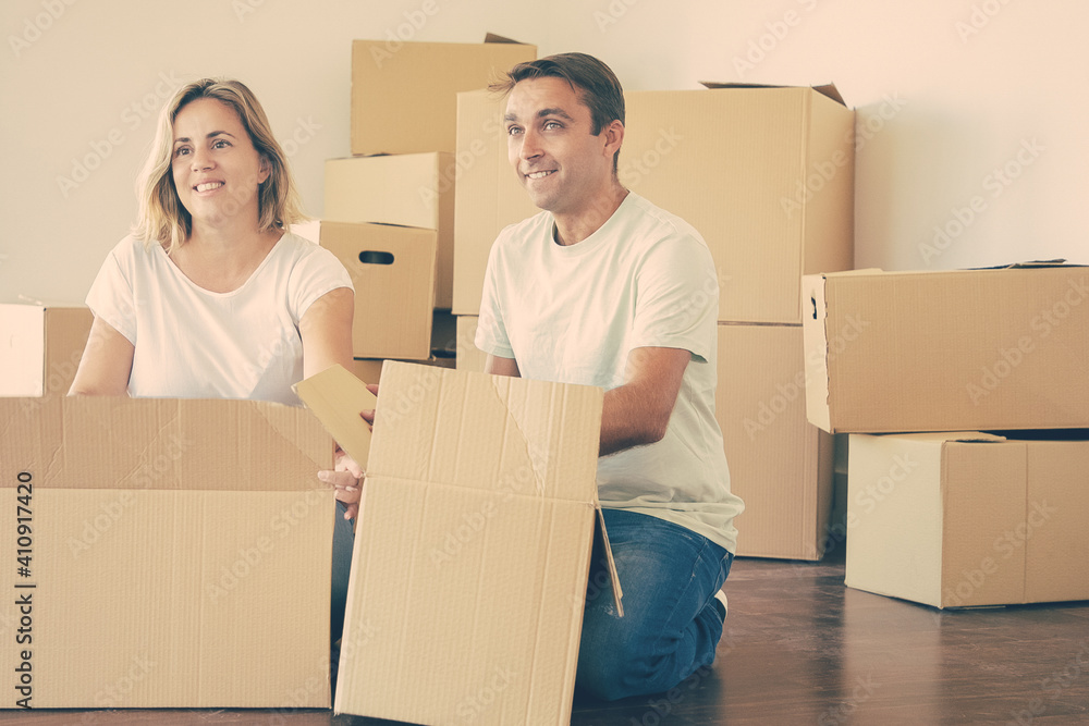 Happy couple unpacking things in new apartment, sitting on floor with open boxes, looking away. Relocation or moving concept Happy couple unpacking things in new apartment, sitting on floor with open boxes, looking away. Relocation or moving concept