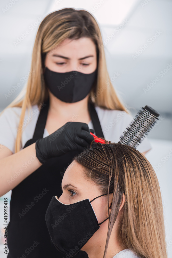 Fototapeta premium Pretty hairdresser with black protective mask and latex gloves - Hairdresser styling a young woman wearing a protective mask.