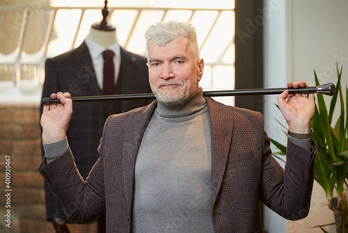 A smiling mature man with gray hair and a sporty physique is holding a fiber carbon cane with two hands behind his head in a clothing store. A male customer with a beard wears a suit in a boutique.