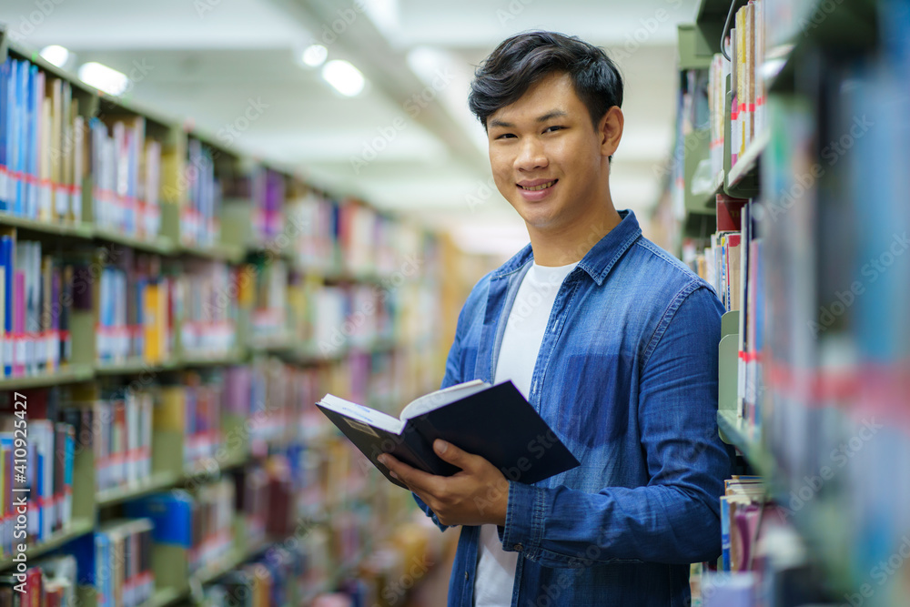 Portrait of Smart Asian man university student reading book and looking at camera between bookshelves in campus library with copyspace.. Portrait of Smart Asian man university student reading book and looking at camera between bookshelves in campus library with copyspace..