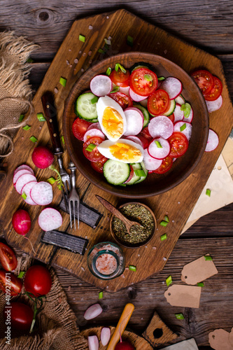 tasty spring salad with round sliced tomatoes, cucumber, radish, boiled eggs, herbs in wooden bowl on rustic table