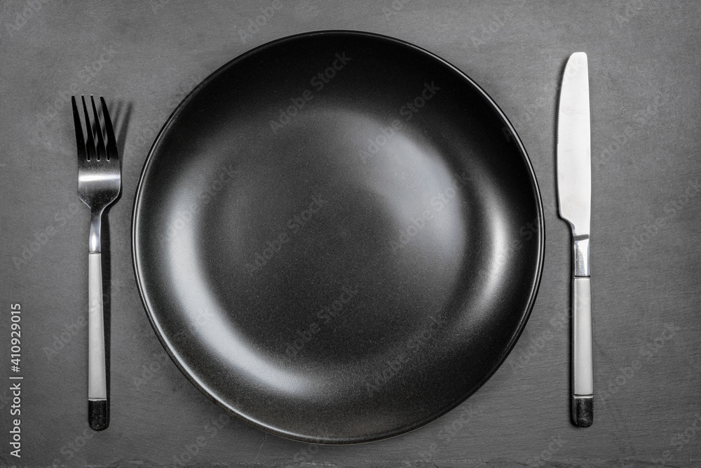 Empty blank black plate with fork and knife on black background of shale serving board. Copy space for prepared dish, recipe or text. Mock up. Top view. Minimal cooking image. Empty blank black plate with fork and knife on black background of shale serving board. Copy space for prepared dish, recipe or text. Mock up. Top view. Minimal cooking image.